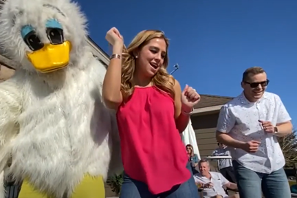 A duck mascot, and young woman and man dancing on outdoor deck 